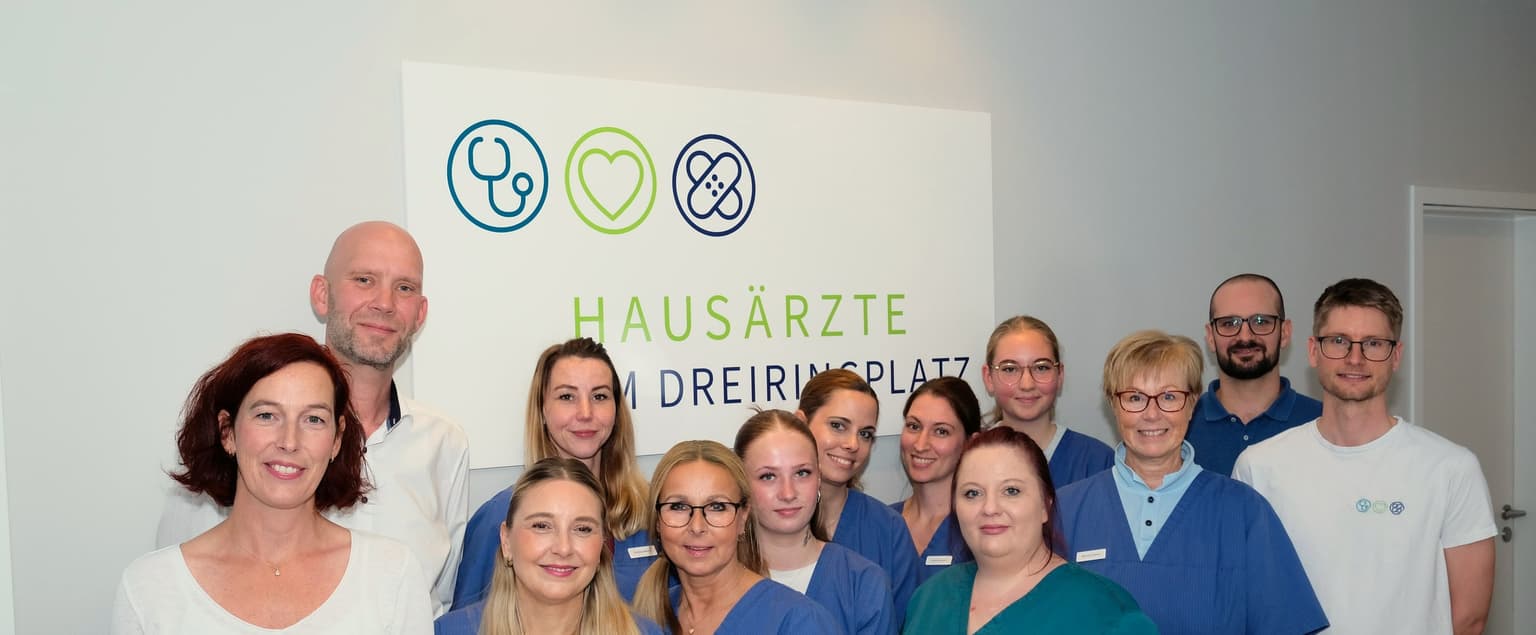Gruppenfoto des gesamten Praxisteams der Hausärzte am Dreiringplatz vor dem Praxisschild